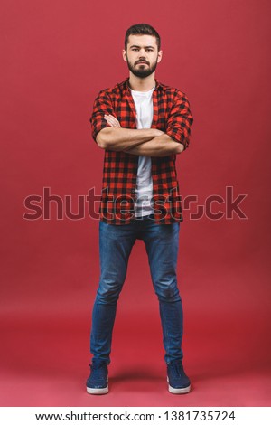 Cool relaxed casual man with crossed arms and titled head looking at camera. Full body length portrait isolated over red background.