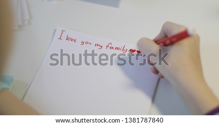 The child writes on a white sheet of paper. Hands girls draw a red heart. I love you my family! 