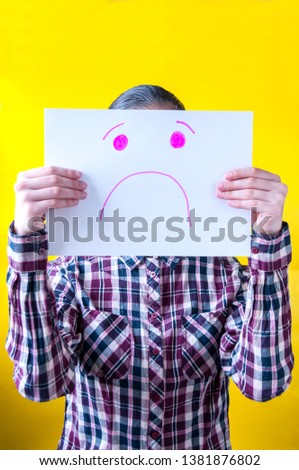 Girl in a plaid shirt on a yellow background holding a white sheet of paper with a painted face of sadness