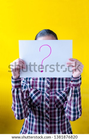Girl in a plaid shirt on a yellow background holding a white sheet of paper with a drawn question mark