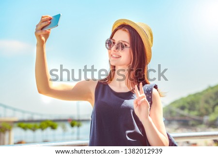 Teenager girl wearing hat and sunglasses standing on the bridge taking selfie photo on sartphone showing v sign smiling cheerful