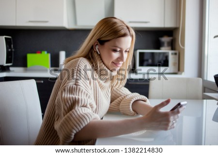 Beautiful young woman in a cozy sweater sitting in the kitchen and listening to music using wireless headphones. Girl at home with phone in hand