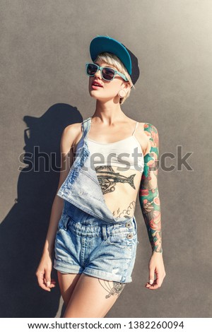 Young alternative girl wearing cap and sunglasses standing isolated on grey wall on the city street looking aside smiling joyful