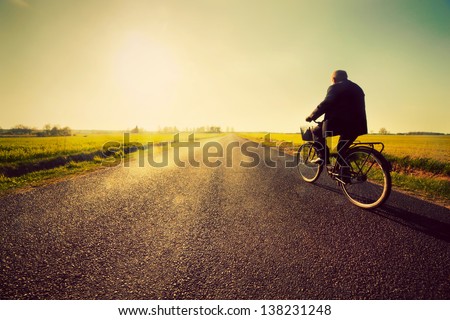 Old man riding a bike on asphalt road towards the sunny sunset sky Royalty-Free Stock Photo #138231248