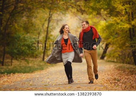 Couple weared in autumn-style clothes (oranfe scarf and vest) running through the autumn landscape. Alley covered with yellow foliage. Autumn walk outdoors. Two lovers in autumn park. Romantic dating Royalty-Free Stock Photo #1382393870