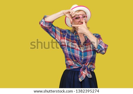 Portrait of attentive modern stylish mature woman in casual style with hat and eyeglasses standing with crop composition gesture and looking at target. indoor studio shot isolated on yellow background