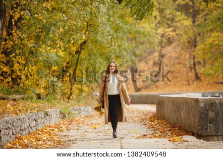 Autumn mood. Romantic woman weared in beige cloak walk in a park on background of beautifull autumn leaves. Smile. fall coming. Autumn story. Brown-haired girl with long hairs. Yellow leaves in hands