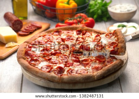 Whole pepperoni pizza pie with a lifted slice of stringy cheesy, on wooden table with ingredients