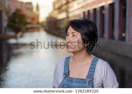 Portrait of a beautiful Asian woman in denim dungarees standing pose near canal and looking afar.