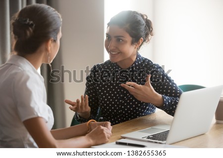 Excited multiethnic female employees discuss work issues sitting at office table, smiling diverse women workers or colleagues engaged in brainstorming talk chatting, explain ideas at workplace Royalty-Free Stock Photo #1382655365