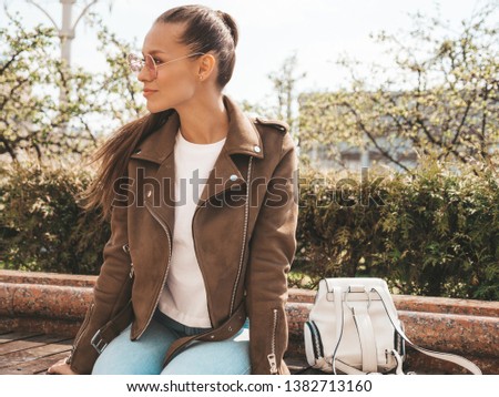 Portrait of beautiful brunette model dressed in summer hipster jacket and jeans clothes. Trendy girl sitting on the bench in the street background. Funny and positive woman in round sunglasses