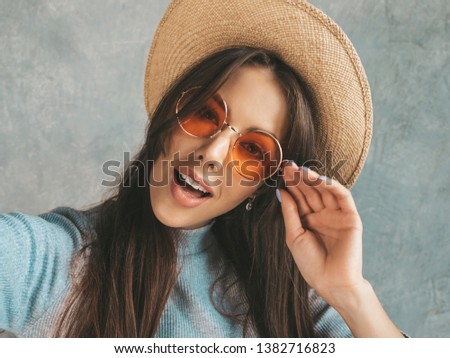Portrait of cheerful young woman taking photo selfie with inspiration and wearing modern clothes and hat. Beautiful girl holding smartphone camera. Smiling model posing near gray wall