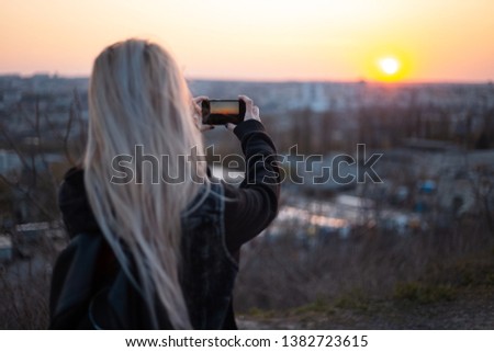 Back view of blonde girl with black backpack, taking photo of sunset near car.