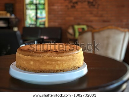Round pound of homemade New York cheesecake on the left hand side of picture in the cafe atmosphere.