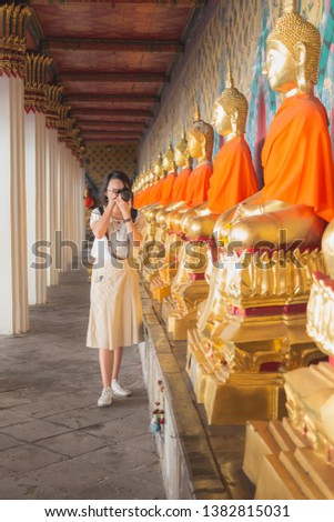 Young Asian woman wearing glasses in white shirt with beige dress carring black bag shooting golden Buddha statues at chuch at Wat Arun, Wat Arun is Temple of Dawn is the favorite historical attract