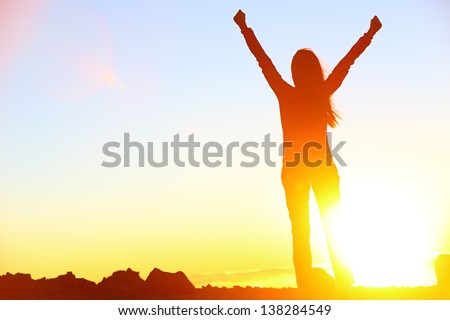 Happy celebrating winning success woman at sunset or sunrise standing elated with arms raised up above her head in celebration of having reached mountain top summit goal during hiking travel trek.