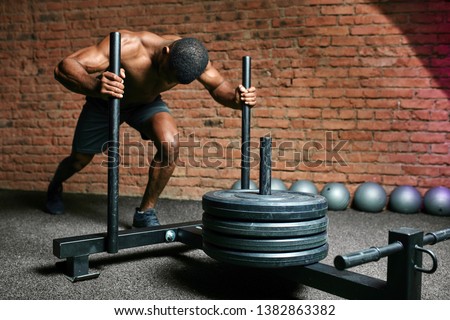 African athletic sportsman pushing sled in cross training gym. Muscular and strong man pushing the sled cross fit equipment on artificial grass turf.