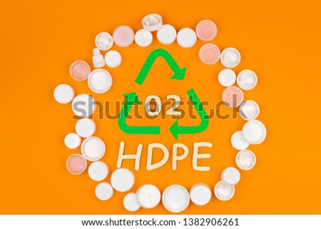 Plastic white reusable hdpe bottle caps stacked in circle around recycling sign symbol with code 02. Nature pollution contamination, ecology environmental problem damage concept. Orange background.