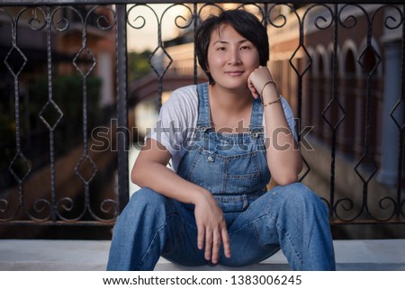Portrait of beautiful Asian woman in denim dungarees sitting pose on a bridge over canal.