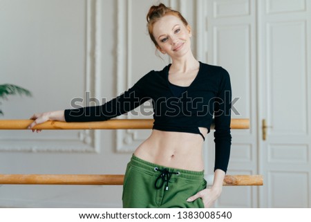 Horizontal shot of satisfied motivated female ballerina wears black top and green trousers, leans hand on ballet barres demonstrates her perfect body, has make up dances in studio has fitness training