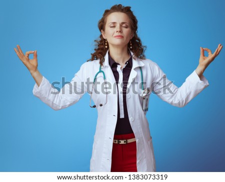 Portrait of relaxed modern physician woman in bue shirt, red pants and white medical robe doing yoga on blue background.