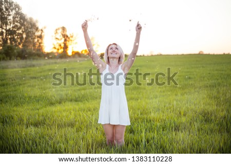 The blonde girl in a tiny white dress with dandelions happily smiles looking up. Green field. Happy moment