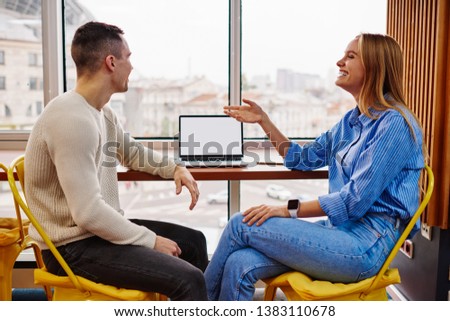 Positive young man and woman dressed in casual wear laughing during friendly conversation at desktop with laptop computer with blank screen area for your internet content.Cheerful students talking