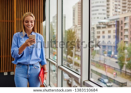 Cheerful female blogger reading funny notification in social networks on modern smartphone device using free 4G internet.Positive young woman chatting online on cellular standing in office interior