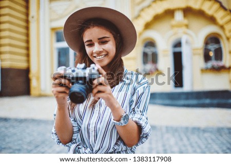 Cheerful caucasian woman in trendy hat viewing picture on vintage camera satisfied with hobby in town, smiling hipster girl holding equipment for taking  photo during  touristic journey on leisure