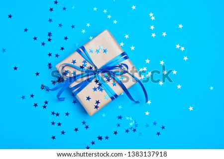 Gift box wrapped in kraft paper with bow and confetti in shape star on blue background, close up