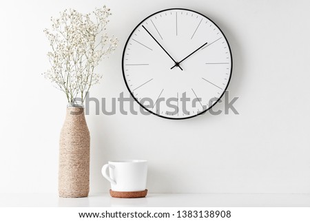 Front view desk with round wall clock, cup and flower in bottle vase on white background. Home office minimal workspace desk