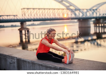 Beautiful Caucasian blonde woman in sportswear stretching while sitting on wall on quay. In background bridge. Push yourself to be great.