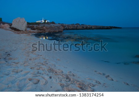 View of white Ayia Thekla chapel above sandy Poseidon Beach near ayia napa, Cyrpus. Blue sky above shallow water with reefs and footprints. Warm evening in fall. Copy space. Long exposure