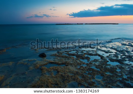 View of empty Poseidon Beach near ayia napa, Cyrpus. Sunset, orange and purple sky among gray clouds above dark blue placid water, shallow water with reefs. Warm evening in fall. Long exposure