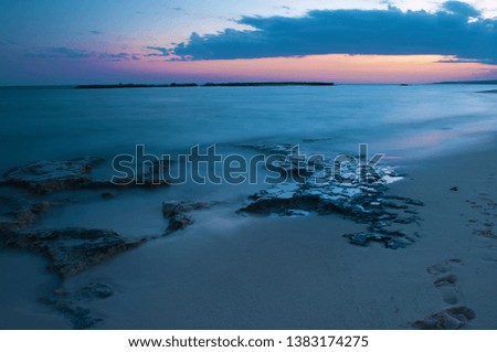 View of empty sandy Poseidon Beach near ayia napa, Cyrpus. Sunset, orange sky, gray clouds above dark blue shallow water, footprints, reefs. Warm evening in fall. Copy space. Long exposure