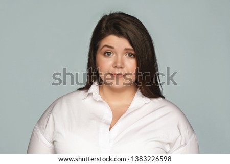 Horizontal image of attractive young overweight plus size female wearing white formal shirt and round earrings posing at blank gray wall, having worried, puzzled or frustrated facial expression