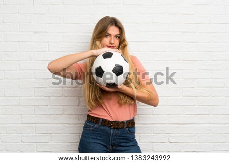 Young blonde woman over white brick wall holding a soccer ball