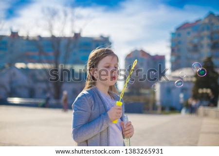 Beautiful girl, has sad face, pretty eyes, brown hair, dressed in white t-shirt and gray jacket, playing soap bubbles in city street. Child portrait. Spring view. Kids fashion style. 