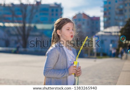 Beautiful girl, has sad face, pretty eyes, brown hair, dressed in white t-shirt and gray jacket, playing soap bubbles in city street. Child portrait. Spring view. Kids fashion style. 