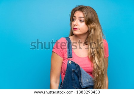 Young blonde woman with overalls over isolated blue wall making doubts gesture looking side