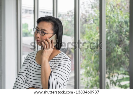 An old Asian woman mother holding a phone in front of a window looking concern and serious.