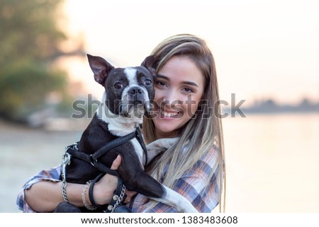Beautiful girl holding on her hands boston terrier black and white dog on sandy beach near the river