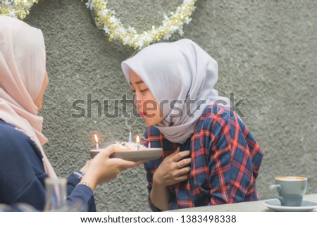 Attractive hijab woman blowing candle from her birthday cake holding by her best friend while sitting on the concrete table at the cafe -image