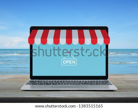 Modern laptop computer with online shopping store graphic and open sign on wooden table over tropical sea and blue sky with white clouds, Business internet shop online concept