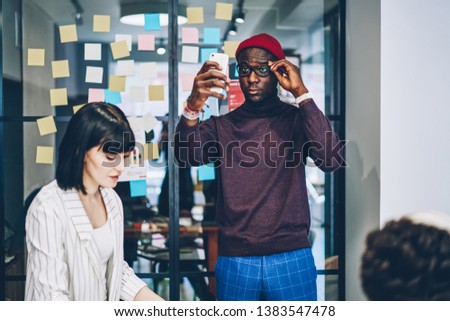 Multiracial male and female colleagues spending time in coworking office during workflow, handsome dark skinned hipster guy in trendy casual wear taking selfie on smartphone camera on work break
