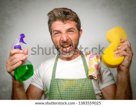 isolated portrait of 30s man in apron holding sponge and detergent spray feeling overwhelmed and bored doing domestic housework of cleaning and washing screaming desperate in stress