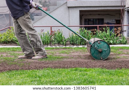 Man in working outfit pulls lawn roller behind. Necessity after long winter and spring for flat surface. Landscaping on the garden. Summer worker Royalty-Free Stock Photo #1383605741