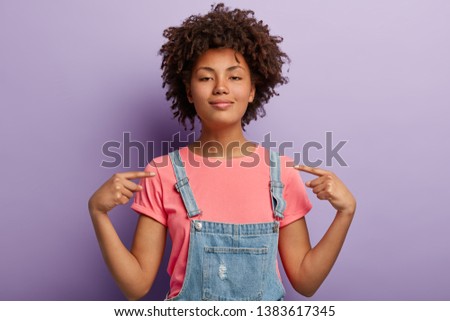 Self assured beautiful African American woman feels proud of her deeds, points at herself feels surge of pride, raises head, has dark healthy skin, wears casual clothes, isolated over purple wall Royalty-Free Stock Photo #1383617345