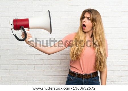 Young blonde woman over white brick wall holding a megaphone