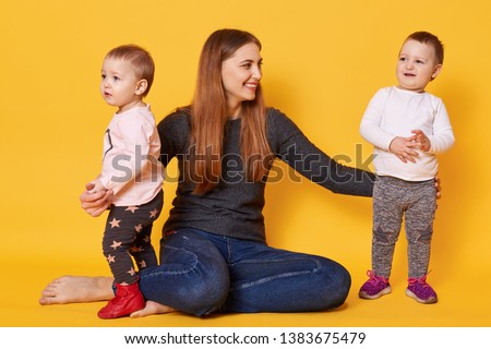 Happy woman, liitle twin girls, mother and her toddlers, try to make photo, infants play with mummy, poses in photo studio isoleted over yellow background. Motherhood, family, children concept. Royalty-Free Stock Photo #1383675479